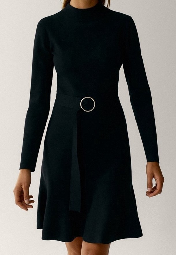 Femme portant une robe noire à manches longues avec un col rond et une ceinture avec une boucle circulaire argentée à la taille.