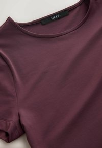 Bordoopunane lühikeste varrukatega t-särk, valmistatud siledast kangast, ümmarguse kaeluse ja minimaalse õmbluse detailidega, musta sildiga, millel on kiri "NEXT".