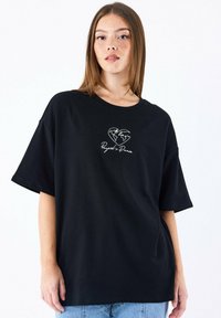 Camiseta negra de corte oversize hecha de algodón, con un gráfico sencillo de un corazón blanco y un globo, acompañado del texto "Project Paris" debajo. Manga corta.