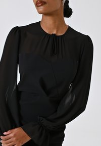 Blouse noire avec des manches longues transparentes, un col rond avec un nœud, et un corsage ajusté au design structuré et à la texture douce.