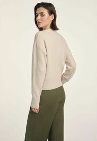 Jasno beżowy sweter z ribbowanym dołem i mankietami, wykonany z miękkiego materiału. W zestawie z ciemnozielonymi spodniami o szerokich nogawkach. Widok z tyłu modelki.