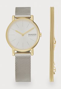 Relógio redondo com um mostrador branco, detalhes em dourado e uma bracelete em malha prateada. Inclui uma pulseira adicional de corrente dourada ao lado.