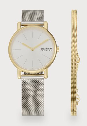 Reloj redondo con una esfera blanca, acentos dorados y una correa de malla plateada. Incluye una pulsera adicional de cadena dorada al lado.