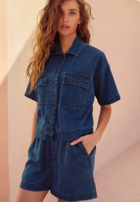 Conjunto de dos piezas de denim que incluye una camisa de manga corta con dos bolsillos en el pecho y un pantalón corto a juego, ambos en color azul oscuro.