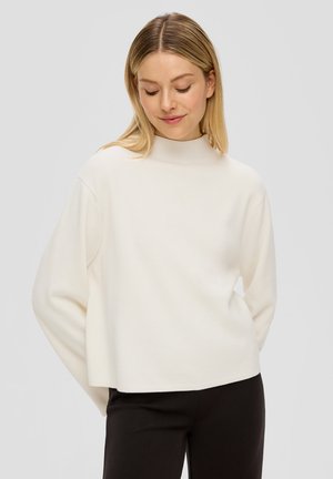 Blonde Frau, die einen lockeren weißen Langarmpullover und schwarze Hosen trägt, mit nach unten gesenktem Blick und den Händen hinter dem Rücken.