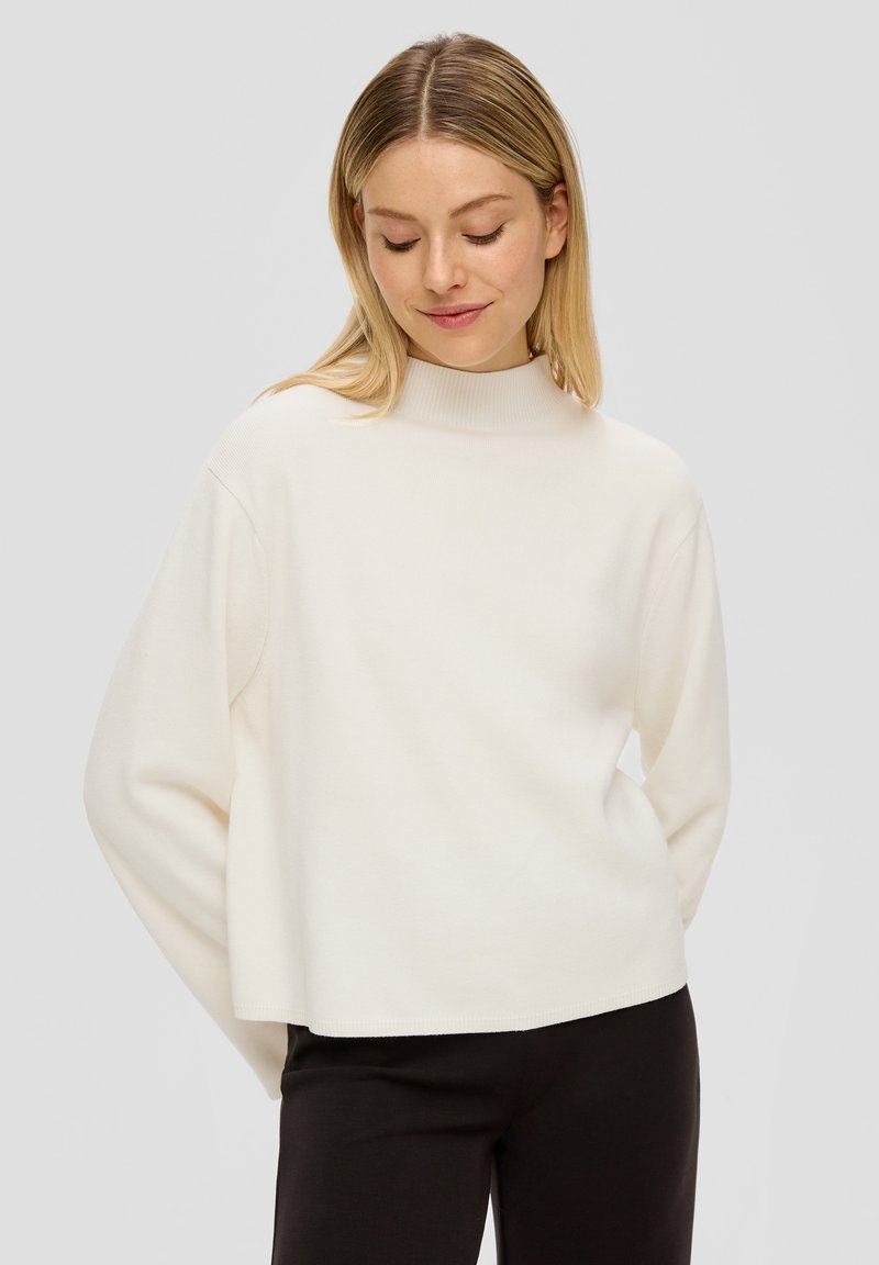 Blonde Frau, die einen lockeren weißen Langarmpullover und schwarze Hosen trägt, mit nach unten gesenktem Blick und den Händen hinter dem Rücken.