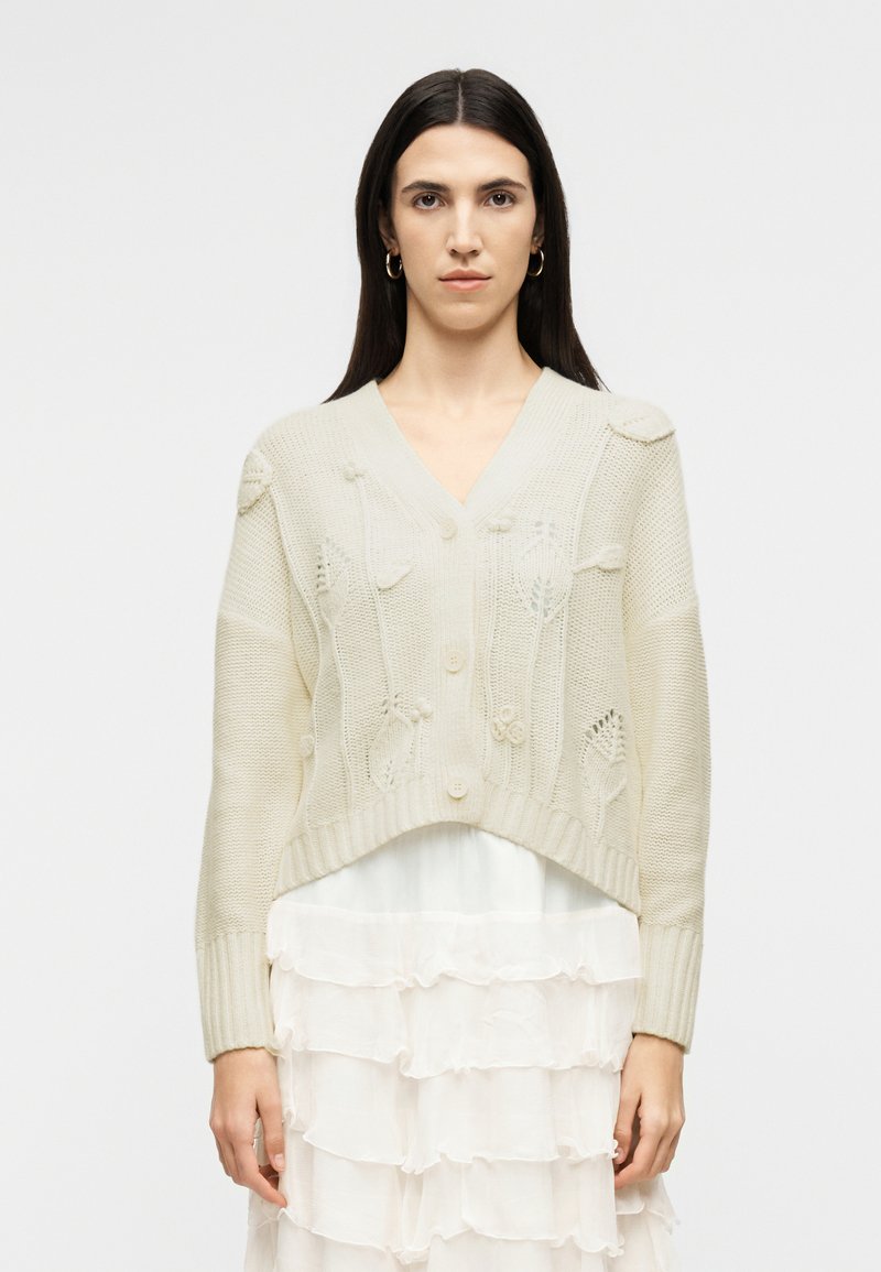 Γυναίκα με μακριά σκούρα μαλλιά φοράει κρεμ πλεκτή ζακέτα με λουλουδένια σχέδια πάνω από λευκό, στρωμένο φόρεμα με βολάν, στέκεται μπροστά σε απλό φόντο.