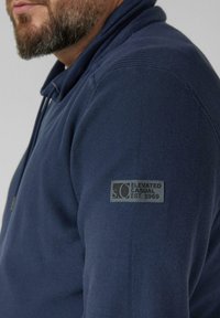 Marineblauer Pullover mit strukturierter Rippoptik und einem Abzeichen am linken Ärmel. Der Kragen ist leicht erhöht gestaltet.