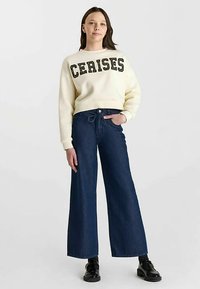 Sweatshirt couleur crème avec "CERISES" en lettres noires, associé à un jean large bleu foncé et des chaussures noires. Texture lisse et coupe décontractée.