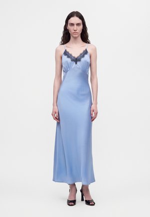 Une femme porte une nuisette bleu clair longue jusqu’au sol, ornée d’une bordure en dentelle foncée, et des sandales noires à talons hauts, sur un fond blanc uni.