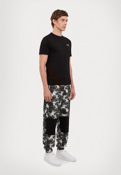 T-shirt en coton noir avec un petit logo doré, associé à un pantalon camouflage vert et blanc avec des patches noirs. Baskets blanches.
