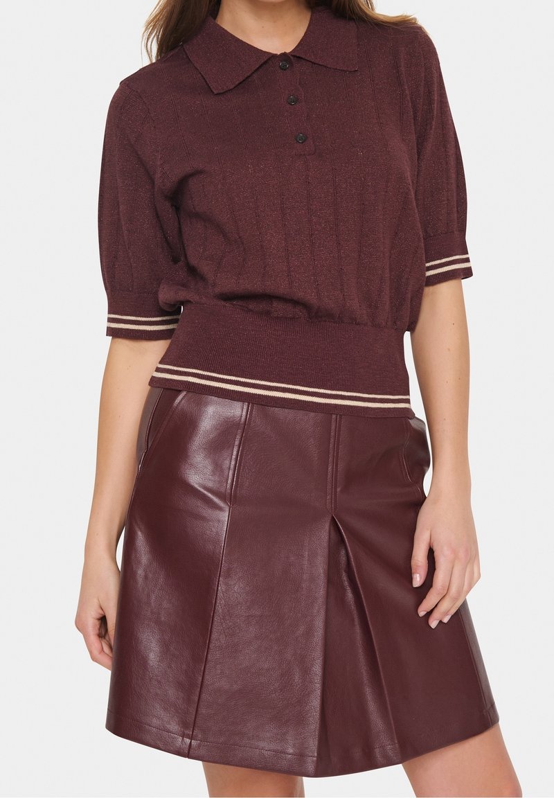 Femme portant un polo en maille bordeaux à manches courtes et une jupe en cuir bordeaux à coupe évasée avec un pli devant, debout devant un fond uni.