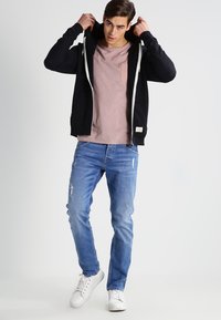Sweat à zip noir avec cordons blancs, t-shirt rose avec poche, jean bleu clair déchiré et baskets blanches. Posture debout.