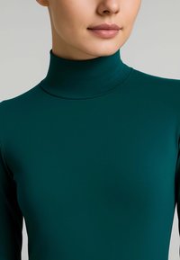 Donna con pelle liscia che indossa un maglione a collo alto fitted di un verde scuro, accorciato sotto il mento e sopra la vita.