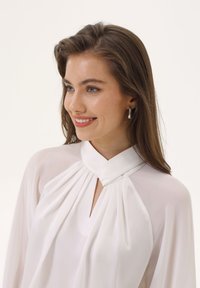 Camicia bianca con collo alto e design plissettato. Presenta un'apertura a goccia sul collo. Tessuto liscio e leggero con una texture morbida.