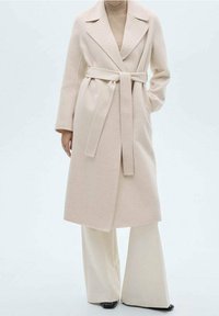 Abrigo de lana beige, con cinturón, cuello de chal y mangas largas; textura suave; combinado con pantalones de pierna ancha color crema y zapatos negros.