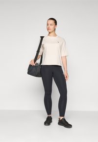 Lichtbeige t-shirt met korte mouwen, zwarte leggings en zwarte sport schoenen. Het model houdt een zwarte schoudertas vast met een tekstuur paneel.