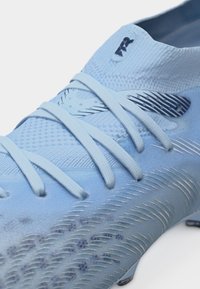 Približen posnetek svetlo modrega nogometnega čevlja z teksturirano zgornjico, križno prepletenimi vezalkami in črnim logotipom blizu ovratnika za gleženj.