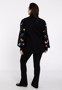 Zwarte oversized sweatshirt met kleurrijke geborduurde ontwerpen op de mouwen, gecombineerd met zwarte flared broek en platte schoenen.