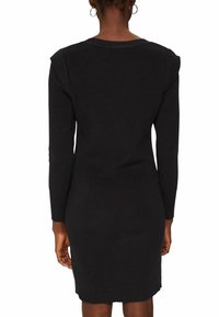 Femme portant une robe en maille côtelée noire à manches longues, vue de dos, avec des boucles d'oreilles créoles et des cheveux courts naturels.