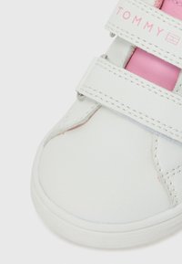Basket blanche avec un accent rose et des sangles en velcro. La chaussure a une texture lisse avec des détails cousus et un logo sur la sangle.