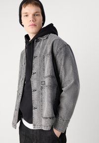 Grå denimjakke med en avslappet passform, med knapper, to fremre lommer og en logo-patch, lagdelt over en svart hettegenser. Teksturert stoff.
