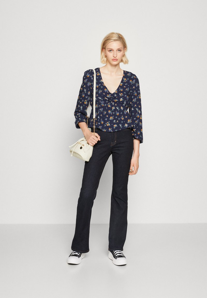 Blusa floreale blu navy con scollo a V profondo e maniche a sbuffo, abbinata a jeans scuri e sneakers bianche, completata da una borsa leggera.