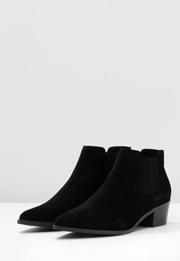 Schwarze Wildleder-Ankelboots mit spitzer Zehenpartie, elastischen Seiteneinsätzen und einem niedrigen, gestapelten Absatz. Glatte Textur mit minimaler Detaillierung.