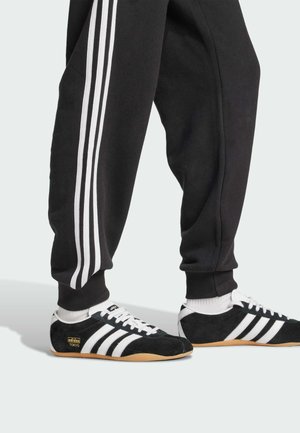 Czarne dresy z trzema białymi paskami po bokach, zestawione z czarnymi zamszowymi sneakerami z białymi paskami i beżową gumową podeszwą.