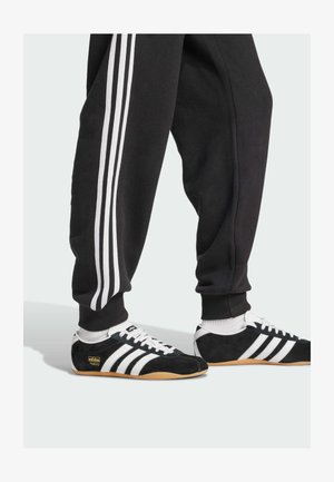 Černé tepláky se třemi bílými pruhy po stranách, doplněné černými semišovými teniskami s bílými pruhy a světlou gumovou podrážkou.