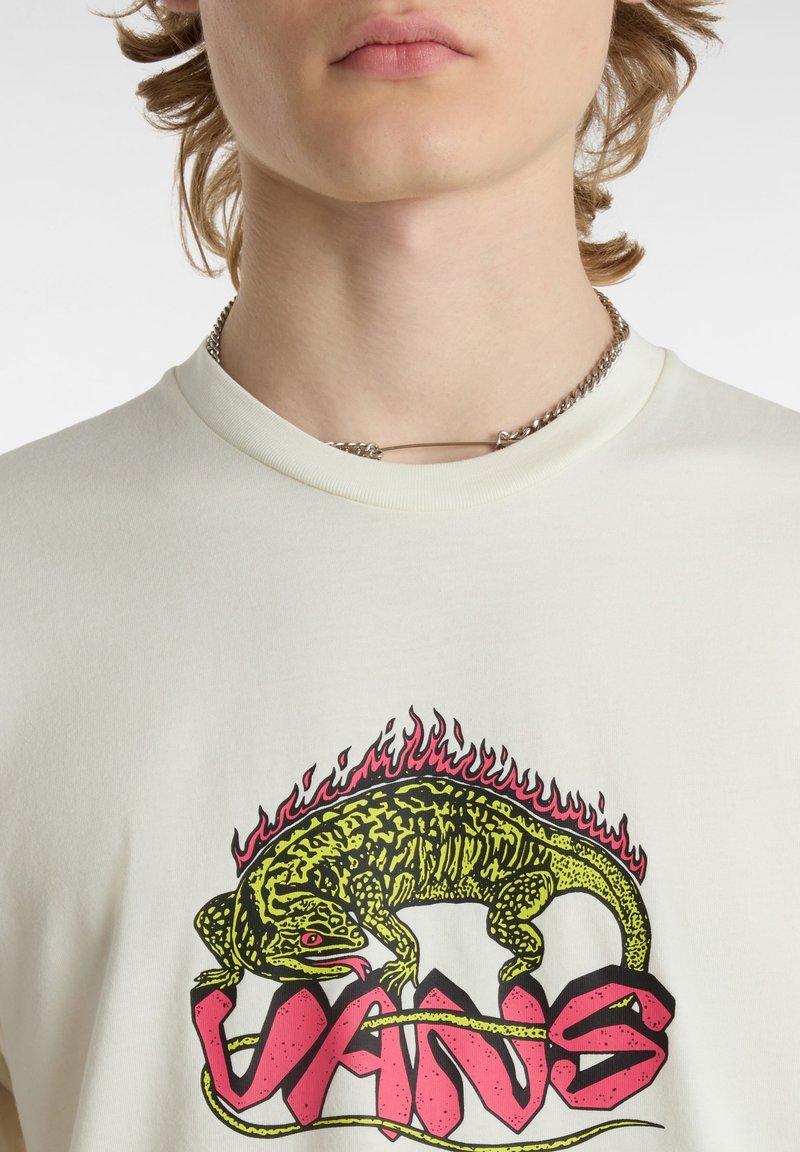 Personne portant un t-shirt blanc avec un lézard vert entouré de flammes au-dessus d'un texte rose stylisé et un collier chaîne argenté fixé avec une épingle.