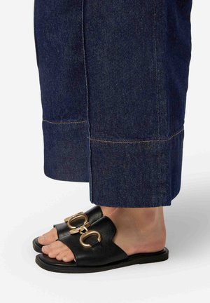 Sandali in pelle nera con una fascia ampia e dettaglio di cinturino circolare dorato, abbinati a pantaloni in denim cropped blu scuro.