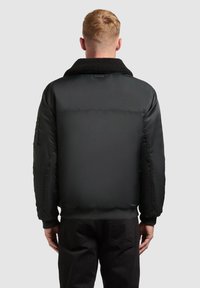 Jachetă bomber neagră cu un guler pufos și texturat, manșete cu nervuri și un finisaj neted, cu închidere cu fermoar și buzunare laterale.