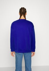 Persona con capelli intrecciati che indossa una felpa a maniche lunghe di un brillante blu e jeans blu, in piedi di fronte a un muro bianco.