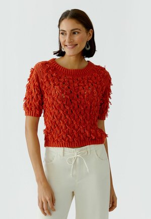 Vrouw glimlachend, draagt een gestructureerde oranje gebreide trui met korte mouwen en witte broek met trekkoord, tegen een effen achtergrond.