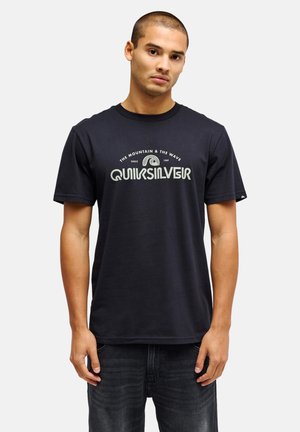 Junger Mann mit kurzen Haaren, der ein schwarzes Quiksilver-T-Shirt und dunkle Jeans trägt, vor einem schlichten hellen Hintergrund stehend.