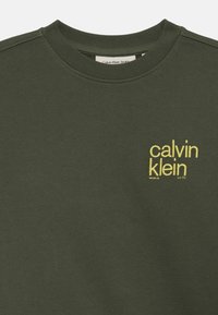 Sweatshirt vert olive en tissu doux et lisse, avec un col rond côtelé et un logo brodé jaune sur le devant.