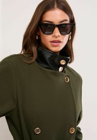 Oliwiona zielona kurtka z wysokim, czarnym, błyszczącym kołnierzem, złotymi guzikami i miękką teksturą. W komplecie z czarnymi, kanciastymi okularami przeciwsłonecznymi.