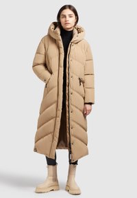 Manteau long beige en duvet avec un motif en chevrons, capuche, fermetures éclair et boutons-pression. Porté par-dessus un haut noir, associé à des bottes couleur crème.