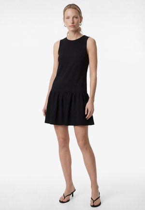 Kvinde stående iført en ærmeløs sort kjole med en rynket nederdel og sorte hælede sandaler mod en ensfarvet hvid baggrund.