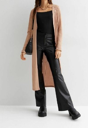 Femme portant un long cardigan beige, un top noir à col carré, un pantalon noir en simili cuir, des bottes noires et portant un sac à main noir.