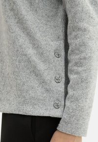 Šedý pletený cardigan s texturovanou tkaninou, postranními knoflíky a rovného střihu. Manžety jsou úzké, což zvyšuje celkový design.