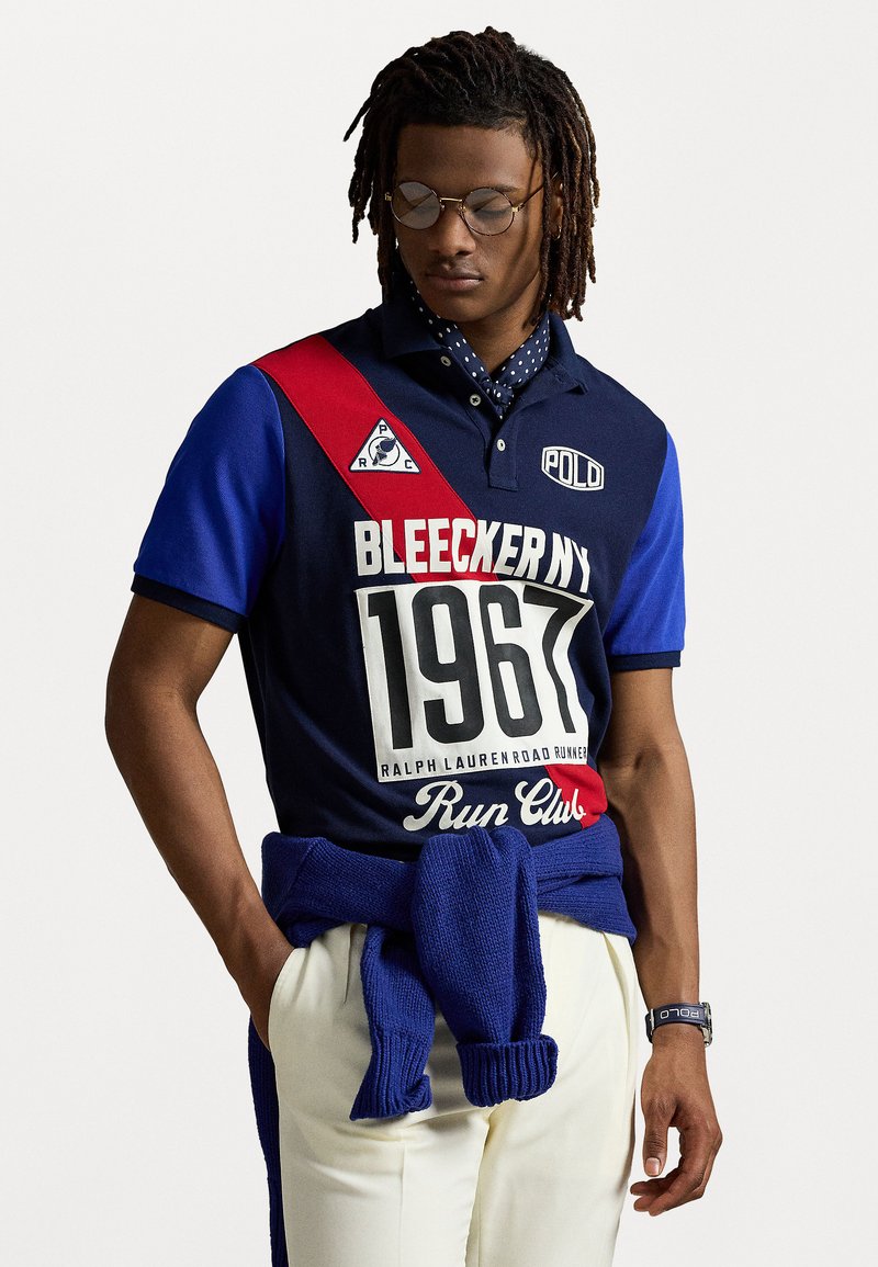 Sötétkék póló piros, fehér és kék díszítésekkel. Feltűnő grafikák, gallér és egy kötött kék pulóver található derékon megkötve.