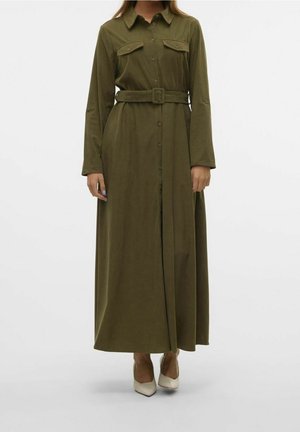 Femme portant une robe longue boutonnée à manches longues vert olive avec des poches poitrine et une ceinture assortie, associée à des talons pointus blancs.