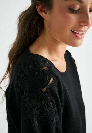 Schwarzer Pullover mit strukturierten, bestickten Details an der linken Schulter und einem Einsatz aus transparentem Stoff. Insgesamt glatter Strick.