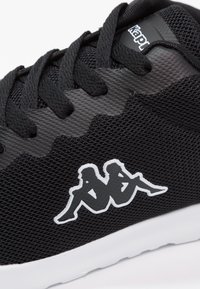 Svart atletisk sko med överdel av mesh, texturerad detaljering, kontrasterande vit sula och en framträdande Kappa-logotyp i vitt på sidan.