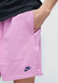 Χέρι στην τσέπη ενός ροζ σορτς Nike που φοριέται με μαύρο πουκάμισο, εμφανίζοντας το λογότυπο της Nike κοντά στο τελείωμα.