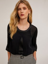 Blusa nera con finitura in satin lucido, scollatura rotonda, piega frontale sottile e maniche a tre quarti, abbinata a pantaloni grigi a fantasia.