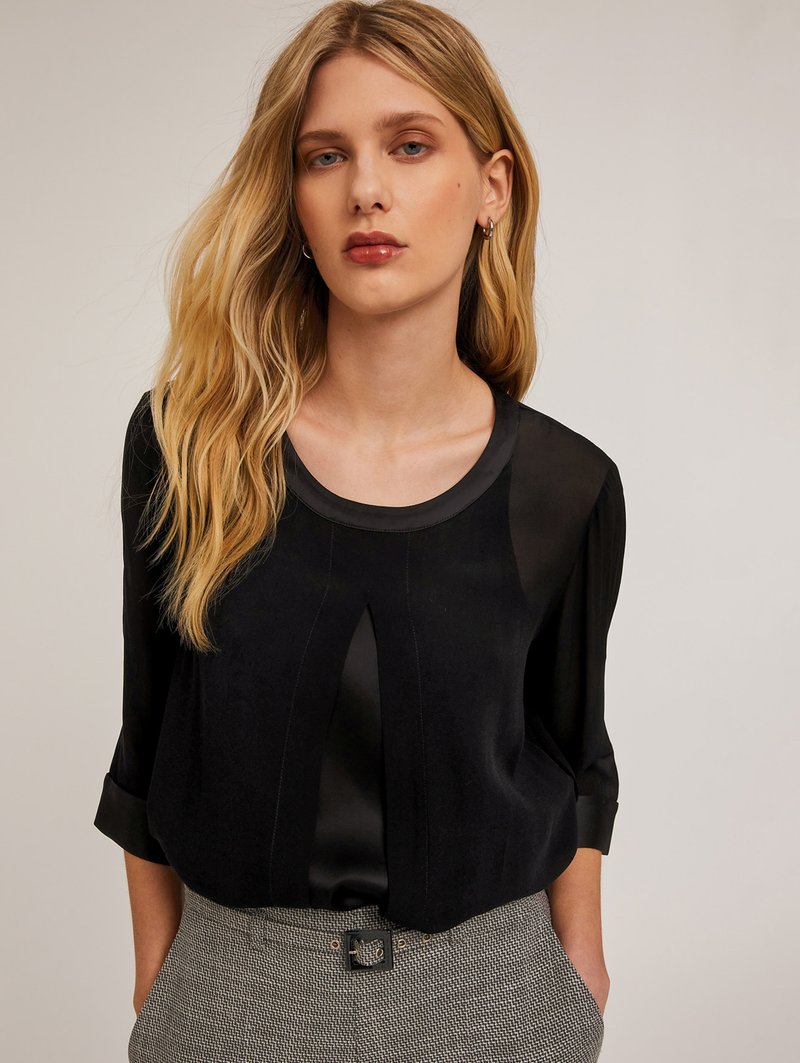 Blusa nera con finitura in satin lucido, scollatura rotonda, piega frontale sottile e maniche a tre quarti, abbinata a pantaloni grigi a fantasia.
