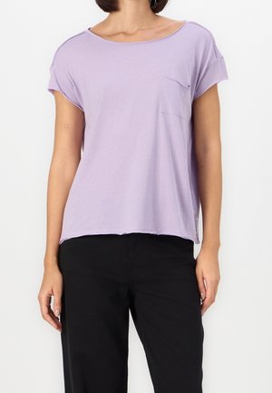 Femme portant un haut lavande à manches courtes et coupe ample avec une poche poitrine, ainsi qu'un pantalon noir, debout devant un fond blanc uni.